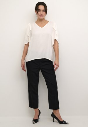 Blouse blanche et transparente avec des manches courtes à volants et un col en V. Associée à un pantalon noir bien taillé et des escarpins noirs pointus. Texture lisse.