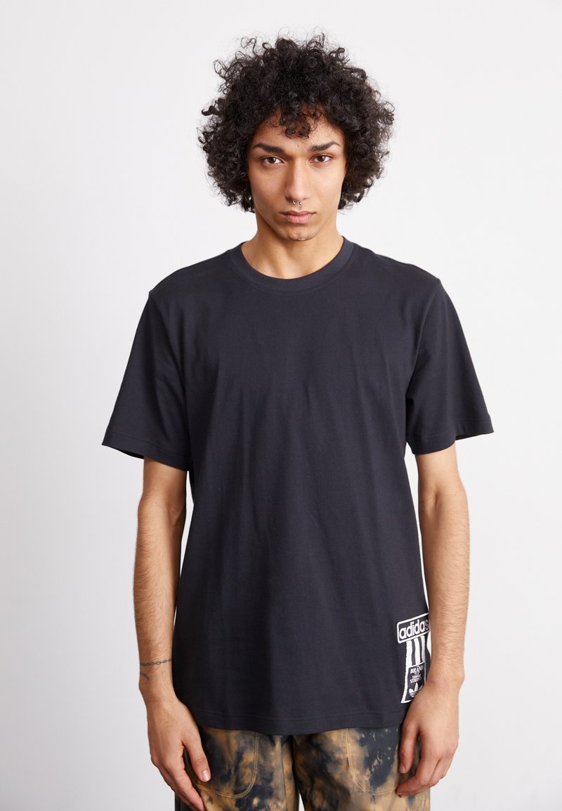 Μαύρο βαμβακερό μπλουζάκι με κοντό μανίκι, στρογγυλή λαιμόκοψη και λευκή ετικέτα λογότυπου στη κάτω αριστερή γωνία, φορεμένο με παντελόνια tie-dye.