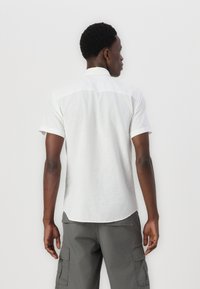 White short-sleeve shirt with a textured fabric and a straight hem. Features a contrasting white collar and cuffs. Back view.