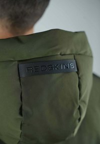 Veste à capuche verte olive en tissu texturé, avec une étiquette logo noire, des coutures lisses et un design de col structuré.
