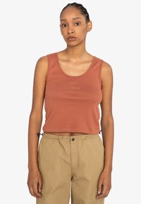 Rustfarvet cropped tanktop lavet af ribbet stof, med rund halsudskæring og broderet logo på fronten. Kombinationen er med lysebrune træk- og slipsebukser.