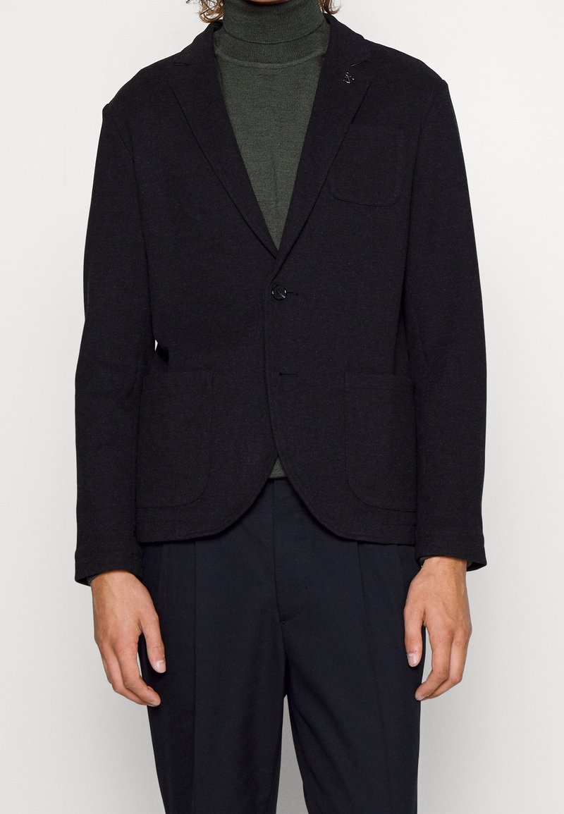 Svart blazer med två framfickor, enkel knappstängning och en något texturerad tyg. Bärs över en mörkgrön polo.