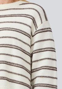 Maglione lavorato a maglia color crema con strisce marrone scuro. Presenta un colletto rotondo e una vestibilità comoda, mettendo in mostra un tessuto testurizzato e cuciture in evidenza.