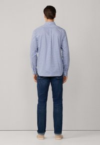 Hombre vestido con una camisa de cuadros azul de manga larga, jeans de color azul oscuro y zapatos beige, de pie frente a un fondo liso gris claro.