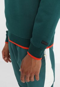 Zielony sweter z pomarańczowymi akcentami przy kołnierzu i mankietach. Żebrowana tekstura oraz czarna metka na mankiecie. W komplecie z pasującymi zielonymi spodniami.