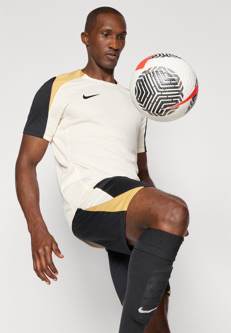 Tenue de sport pour homme comprenant une chemise claire avec des accents noirs et dorés, un short noir et des chaussettes noires avec un logo blanc.