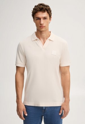 Jeune homme aux cheveux bouclés portant un polo beige clair à manches courtes et un jean bleu, debout devant un fond uni.