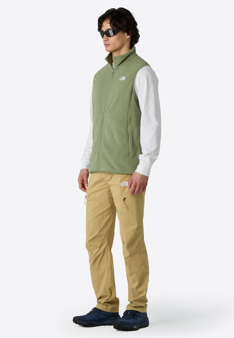 Grüne Fleece-Weste mit hohem Kragen, Reißverschluss vorne und Seitentaschen, kombiniert mit beigen Hosen und blauen Wanderschuhen. Schwarze Sonnenbrille.