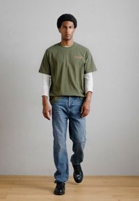 T-shirt de algodão verde-oliva sobre uma camisa de manga longa branca, combinada com jeans de denim azul claro e sapatos pretos, a caminhar sobre um piso de madeira.