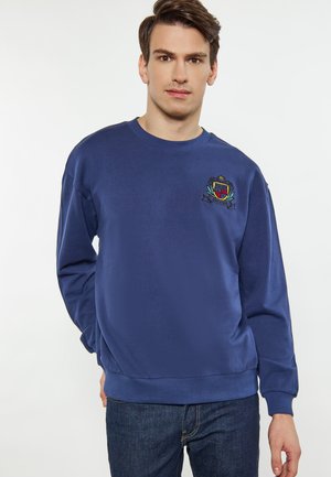 Mørkeblå langærmet sweatshirt lavet af blødt stof, med et broderet emblem på brystet og ribbede manchetter og kant.