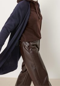 Navy gerippter Cardigan, brauer Hoodie und glänzende braune Lederhosen. Merkmale sind Kordeln und Taschen an der Hose.