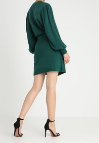 Robe verte en maille avec manches longues bouffantes, taille cintrée et ourlet légèrement asymétrique. Une paire d'escarpins noirs à brides complète la tenue.