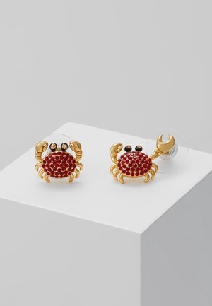 Boucles d'oreilles clous en forme de crabe en or avec corps en pierres précieuses rouges et yeux noirs, présentées sur un socle blanc contre un fond gris.