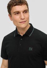 Czarna koszulka polo z krótkim rękawem, z szarym wykończeniem kołnierzyka i trzema guzikami. Po lewej stronie na piersi subtelne logo. Gładka tekstura.
