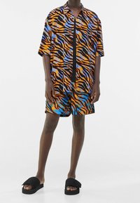 Koszula z krótkim rękawem na guziki oraz pasujące krótkie spodenki z wzorem w paski tygrysa w kolorach pomarańczowym, niebieskim i czarnym, zestawione z czarnymi klapkami.
