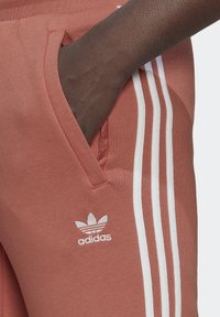 Adidas sweatpants in rust color, featuring three white stripes along the side, cotton material, and a front pocket with logo detail.