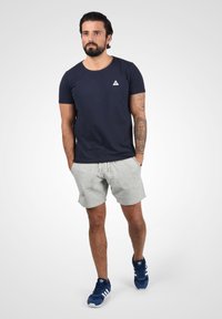 Granatowa bawełniana koszulka z małym logo w kształcie trójkąta, szare szorty z kieszeniami oraz niebieskie sneakersy z białymi akcentami. Projekt stroju casual.