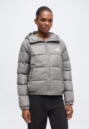 Joven mujer con chaqueta acolchada gris de The North Face con capucha y pantalones negros, de pie frente a un fondo liso y claro.