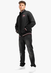 Zwarte zip-up jas met een witte en rode gestreepte kraag en manchetten, gecombineerd met donkere jeans en witte sneakers. Heeft voorzakken en logo-detail.