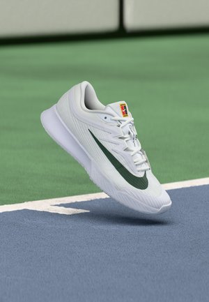Zapatilla deportiva blanca con el logo de Nike en verde oscuro flotando sobre líneas azul y verde de una cancha de tenis.