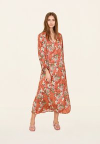 Robe longue et fluide en rouille, ornée de motifs floraux en blanc, vert et jaune. Décolleté en V, manches longues, fabriquée en tissu léger. Chaussures à talons carrés transparentes.