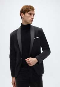 Schwarzer Samtblazer mit Satinrevers, Ein-Knopf-Verschluss und einer paspelierten Taschenverzierung. Dazu ein schwarzer Rollkragenpullover. Glatte Textur.