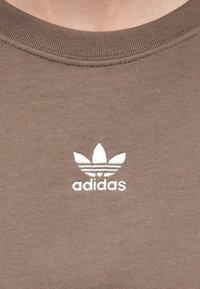 Sweatshirt din bumbac maro, cu un logo alb Adidas și trei dungi asemănătoare frunzelor deasupra numelui mărcii. Gât rotund și guler ribbit.