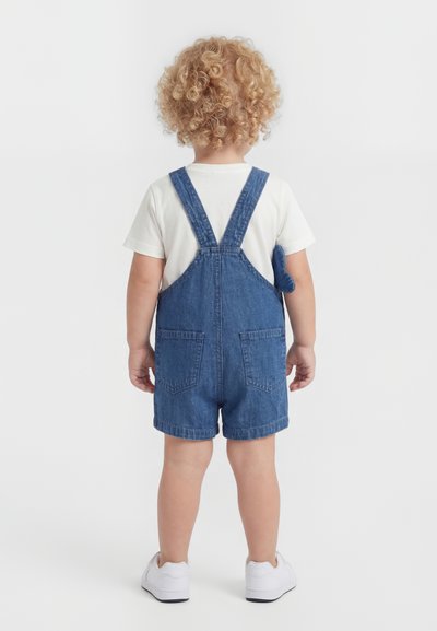 Νήπιο με σγουρά ξανθά μαλλιά που φοράει λευκό μπλουζάκι, μπλε τζιν κοντό σαλοπέτα και λευκά αθλητικά παπούτσια, στέκεται αντικρίζοντας ένα λευκό φόντο.