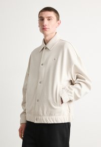 Giubbotto bomber beige con chiusura a bottoni, tasche laterali e fondo a coste. Presenta un piccolo logo ricamato sul lato sinistro del petto.