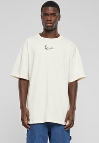 Krämfärgad oversized t-shirt i bomull, med en svart handskriven logga på bröstet, korta ärmar och en avslappnad passform.