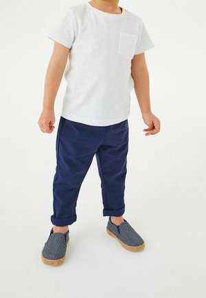 Kind trägt ein weißes Kurzarmhemd, marineblaue hochgekrempelte Hosen und blaue Slip-on-Schuhe mit Streifen und steht vor einem weißen Hintergrund.