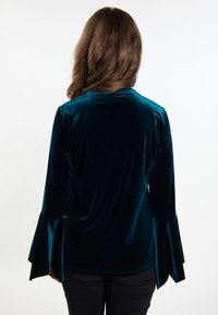Top in velluto teal con maniche lunghe e svasate e colletto rotondo. Il tessuto ha una superficie liscia e una leggera lucentezza.