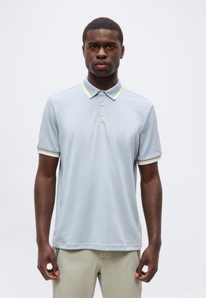 Ung mand stående iført en lyseblå kortærmet polo med hvidstribet krave og beige bukser, neutralt baggrund.