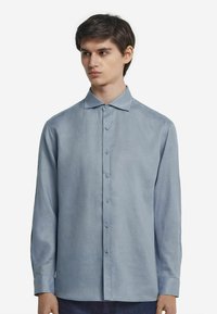 Ljusblå långärmad skjorta med krage, knappfram, och ett glättat, något texturerat tyg. Knapparna matchar skjortans färg.