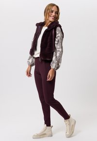 Faux fur vest in dieppaars, gedragen over een top met een patroon met lange mouwen en gecombineerd met bordeaux leggings en beige hoge sneakers.