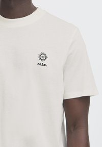 Krämfärgad bomullströja med korta ärmar, som har en svart solgrafik och texten "calm." broderad på vänster bröst. Slät textur.