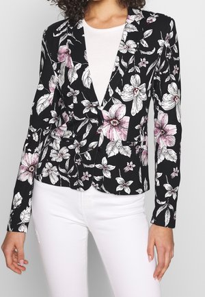 Femme portant un blazer noir avec un motif floral rose et blanc par-dessus un haut blanc et un pantalon blanc, debout devant un fond neutre.