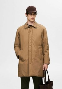 Cappotto marrone, lungo fino al ginocchio, con taglio dritto, chiusura a bottoni e tasche laterali. Indossato sopra una camicia scura e abbinato a un cappellino da baseball scuro.