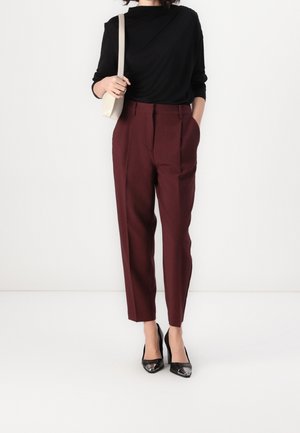 Femme portant un haut noir à manches longues, un pantalon bordeaux sur mesure, des talons en cuir verni noir et portant un petit sac à épaule beige.