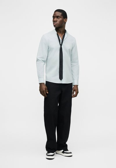 Άνδρας που φορά ανοιχτό γκρι πουκάμισο με κουμπιά, με χαλαρό μαύρο γραβάτα, μαύρο παντελόνι και ασπρόμαυρα αθλητικά παπούτσια, στέκεται μπροστά σε απλό φόντο.