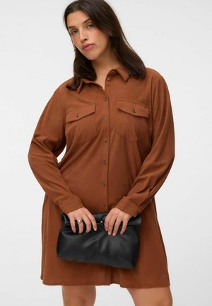 Woman with dark hair wearing a brown button-up dress holding a black folded clutch bag against a white background.