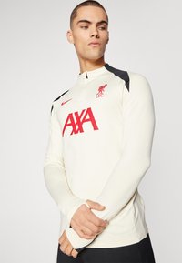 Długi rękaw sportowej koszulki w kolorze kremowym z czarnymi akcentami, z wysokim kołnierzem, zamkiem błyskawicznym, czerwonym logo Liverpool FC oraz napisem "AXA".
