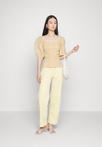 Top amarelo xadrez com mangas bufantes e decote quadrado, combinado com calças amarelas claras de perna reta e sandálias brancas de tiras, segurando uma mala branca.