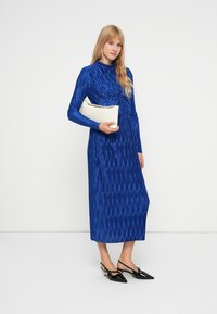 Mėlyna ilgomis rankovėmis suknelė su tekstūrinėmis vertikaliomis linijomis, prigludusiu liemeniu ir plazdančia sijona, derinama su kremine rankine ir juodais smailiais bateliais.