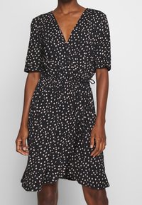 Robe noire cintrée avec des manches courtes, présentant un décolleté en V et une taille cintrée. Elle a un motif à pois rose clair sur un fond sombre.