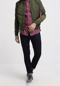Uomo che indossa una giacca bomber verde oliva, una camicia a quadri rossi, jeans scuri e sneakers grigie mentre cammina su uno sfondo bianco uniforme.