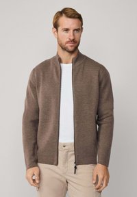 Hnedý zipsový sveter s vysokým golierom, vyrobený z mäkkého materiálu. Má rovný strih a dlhé rukávy. Nosený cez biely tričko.