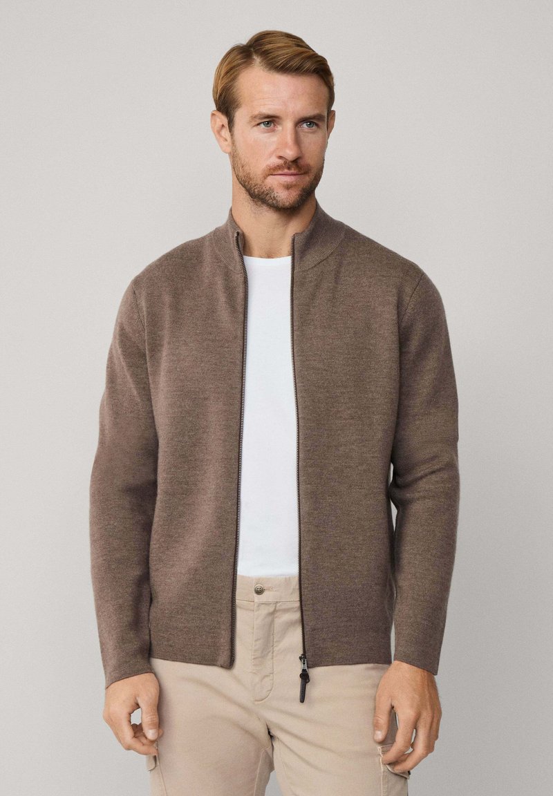 Hnedý zipsový sveter s vysokým golierom, vyrobený z mäkkého materiálu. Má rovný strih a dlhé rukávy. Nosený cez biely tričko.