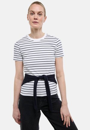 Femme portant une chemise blanche à manches courtes avec des rayures horizontales noires, un pantalon noir et un pull noir noué autour de la taille.