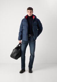 Marineblå pufferjakke med rød foring, sort turtleneck, mørke jeans og sorte støvler. Bærer en sort læder sportstaske.